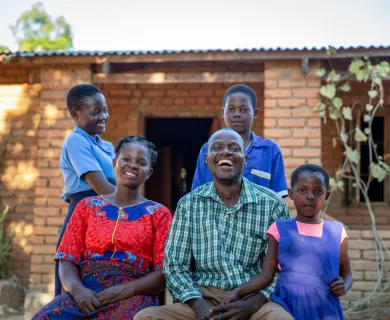 A man and woman, sitting next to each other, smile and laugh. They are surrounded by their three kids, one of whom is a young girl clutching her father's hand.